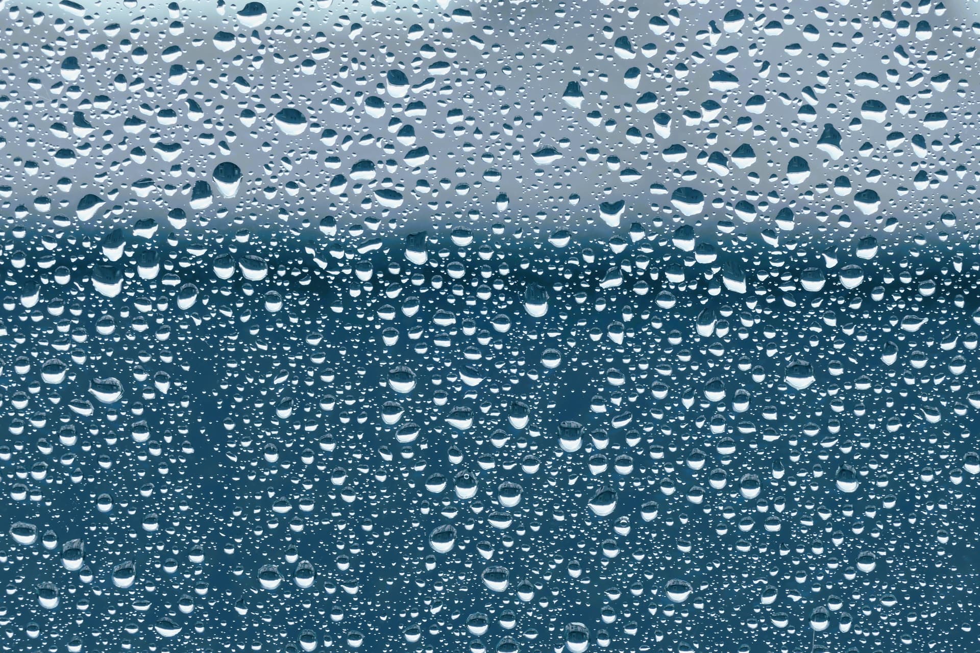 Water beaded across a pane of window glass during rain, with the view behind it blurred.